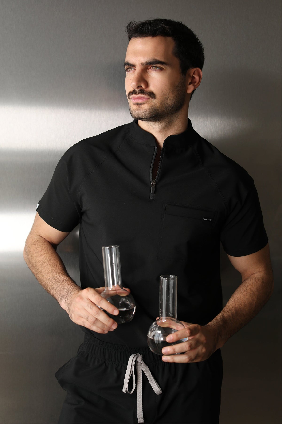 Man wearing the Harry Black W&C Shield™ scrub top while holding two laboratory flasks filled with clear liquid.